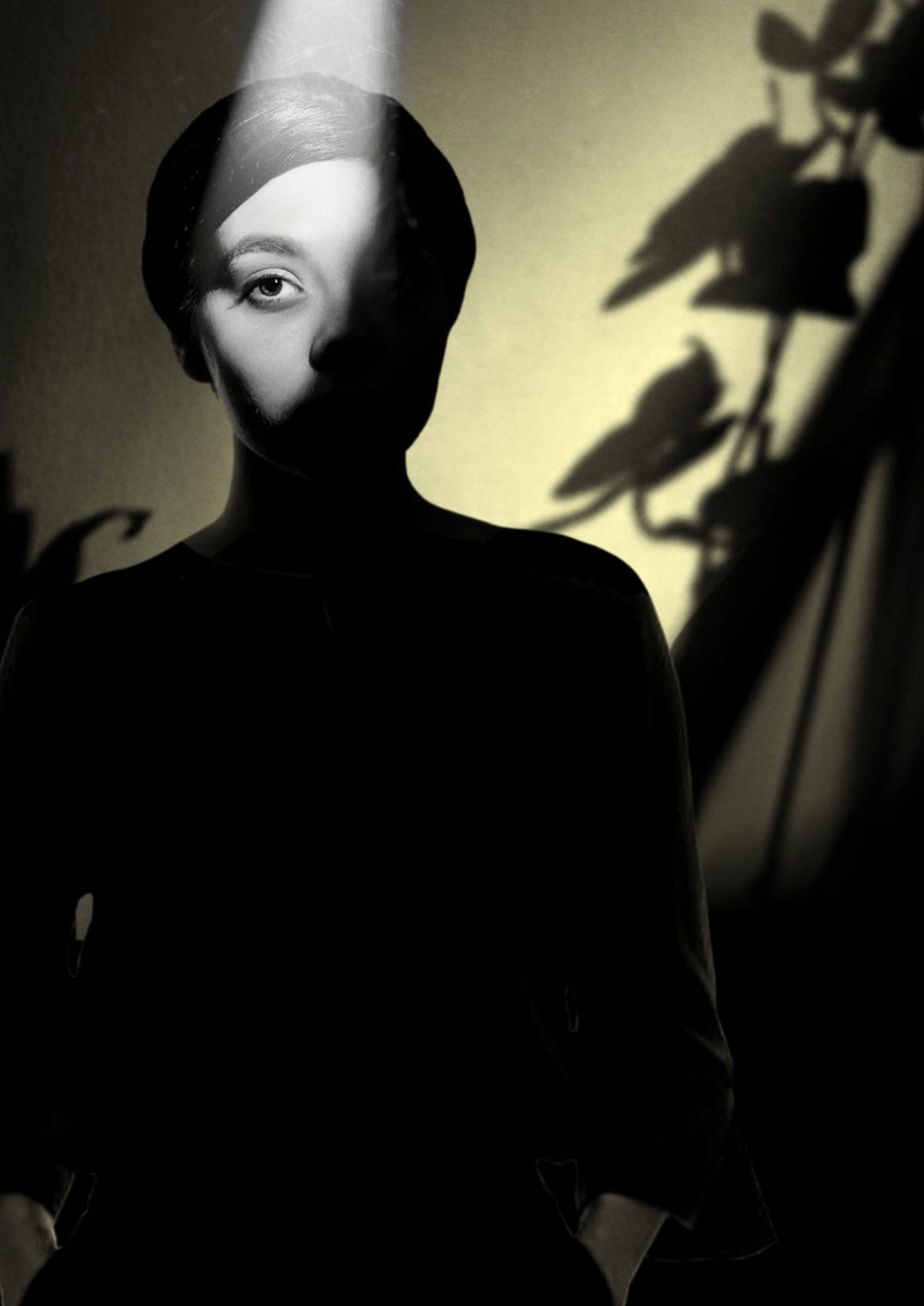 A woman half-hidden in shadow. A single beam of light falls from above and illuminates one eye. A dramatic black-and-white photograph against a warm sepia wall.